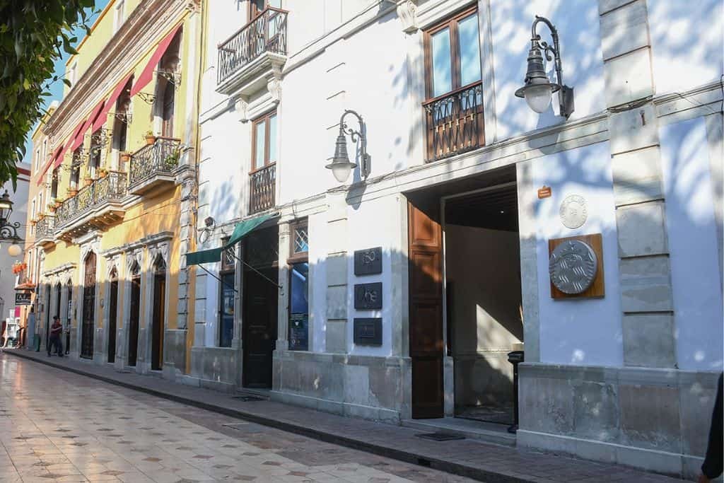 A cobblestone street lined with colorful colonial buildings features wrought-iron balconies and decorative wall lamps. Sunlight casts tree shadows across the white and yellow facades.