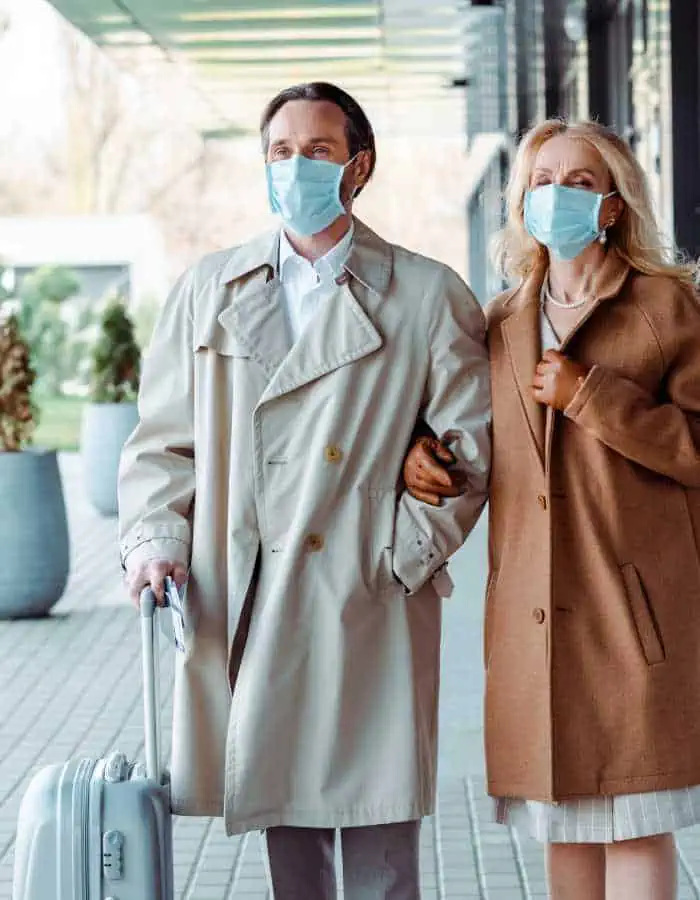 This image shows a man and woman wearing face masks, walking arm in arm outside a modern building. The man is pulling a rolling suitcase, suggesting they are traveling.
