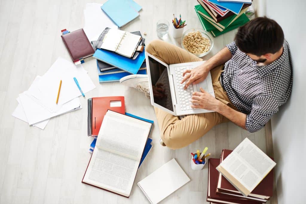 Man sitting on the floor with a laptop and books spread around him researching how to start a travel blog without traveling.