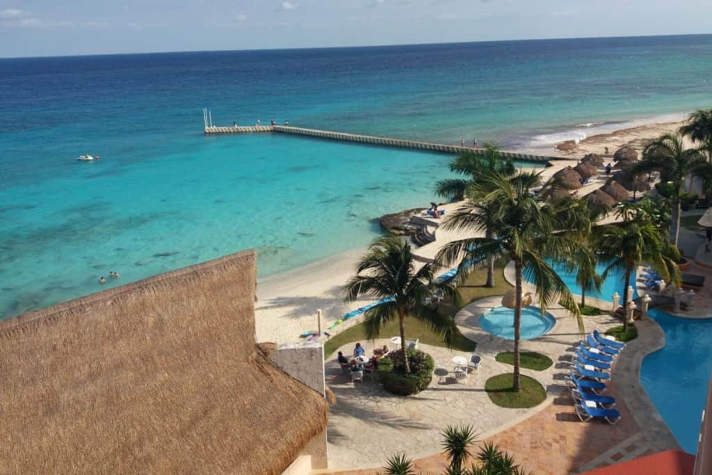 Beachfront resort features turquoise water, a long pier extending into the sea, and palapa-covered lounge areas along the sand. Multiple pools with palm trees and blue lounge chairs create a tropical vacation setting.