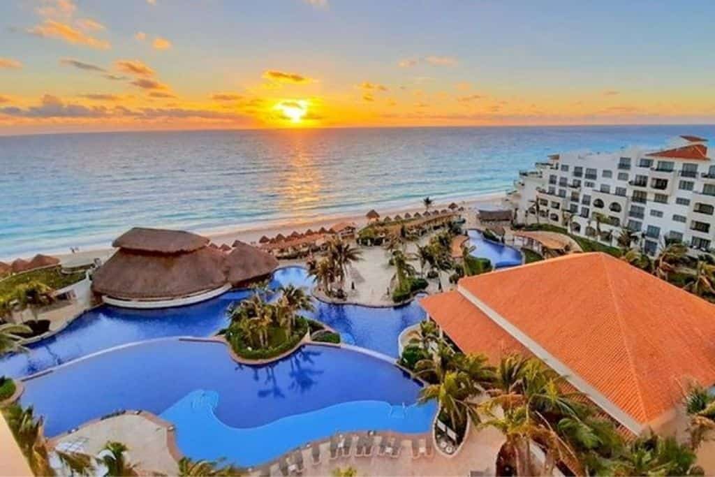 A resort overlooking the ocean at sunrise, with the sun casting golden light over the water. The property features multiple pools with curving edges, palm trees, and thatched-roof palapas. White buildings with balconies face the beach, enhancing the tropical luxury setting.