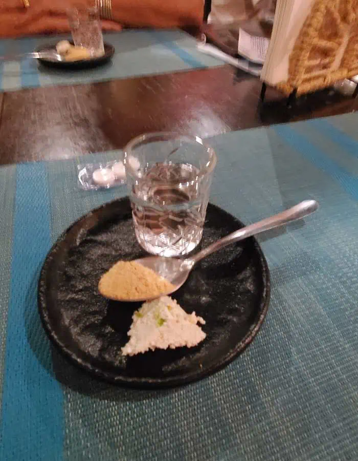 A glass of mezcal placed on a small black plate, accompanied by a spoon holding sugar and a bit of soft cheese. The setup is on a blue woven tablecloth, indicating a casual yet refined tasting experience.