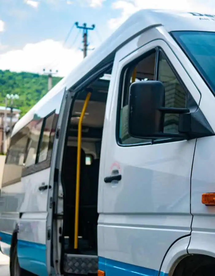A small white tour bus with its door open, ready for passengers to board. The bus has a blue stripe along its lower edge and is parked outdoors, with a glimpse of greenery and a partly cloudy sky in the background.