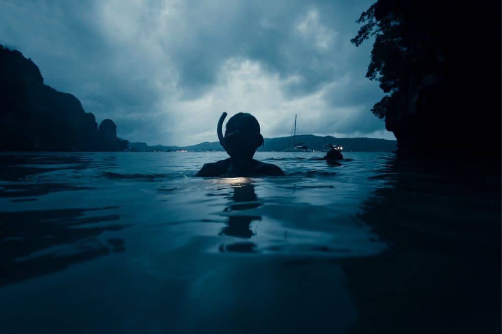 Silhouetted snorkeler emerging from dark waters near a cave or rocky coastline at dusk or dawn, with dramatic cloudy skies and distant landforms in the background.