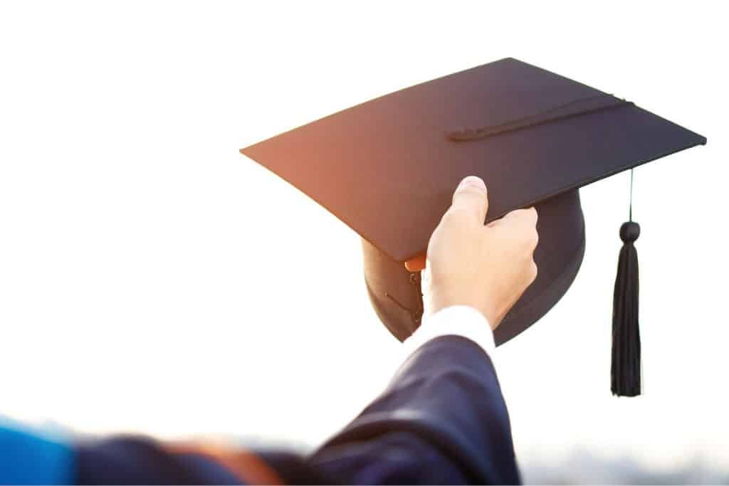 This image features a graduate holding up a black mortarboard cap with a tassel against a bright, sunlit background. The person is dressed in a traditional graduation gown, symbolizing achievement and academic success. The scene conveys a sense of accomplishment, new beginnings, and the excitement of future opportunities.