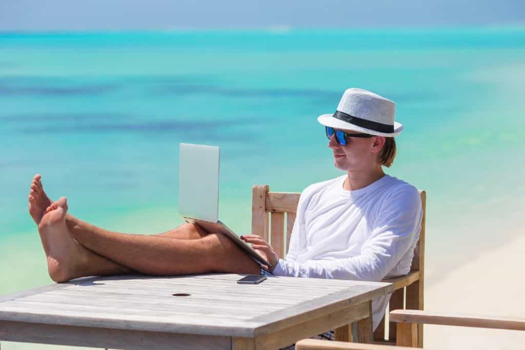 This image features a relaxed man working on his laptop at a wooden table on a pristine beach. He is dressed in a white long-sleeve shirt, a stylish fedora, and sunglasses, with his feet propped up, enjoying the turquoise ocean view. The scene embodies the digital nomad lifestyle, combining work with tropical leisure.