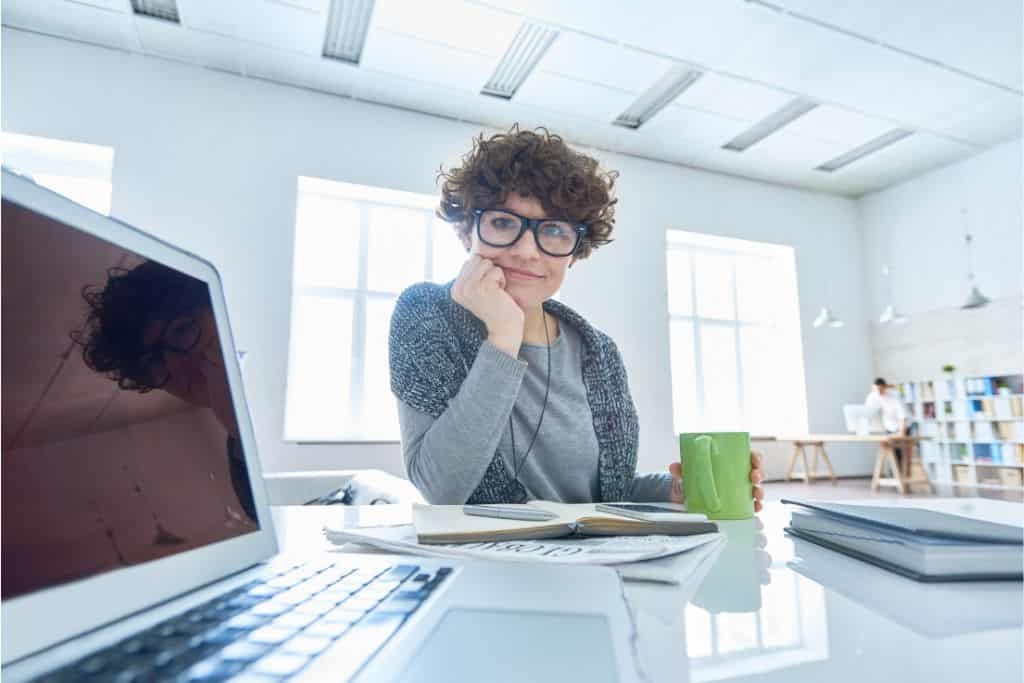 This image features a woman with curly hair and glasses sitting at a modern, bright workspace. She is smiling while holding a green coffee mug, with an open laptop and notebooks in front of her. Large windows flood the room with natural light, creating a productive and inviting atmosphere.