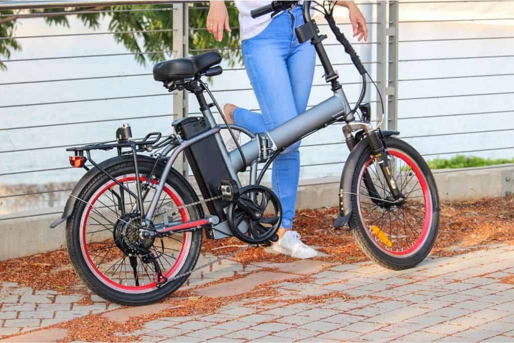 Person standing beside a modern black electric bike with red-rimmed wheels, parked on a brick pathway near a metal railing.