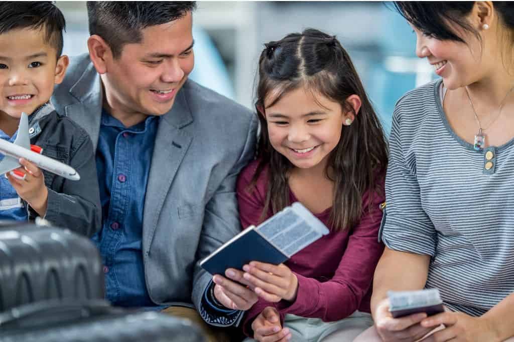 Smiling family sits together holding passports and boarding passes, likely waiting at an airport. A young boy holds a toy airplane while his sister and parents look at travel documents.