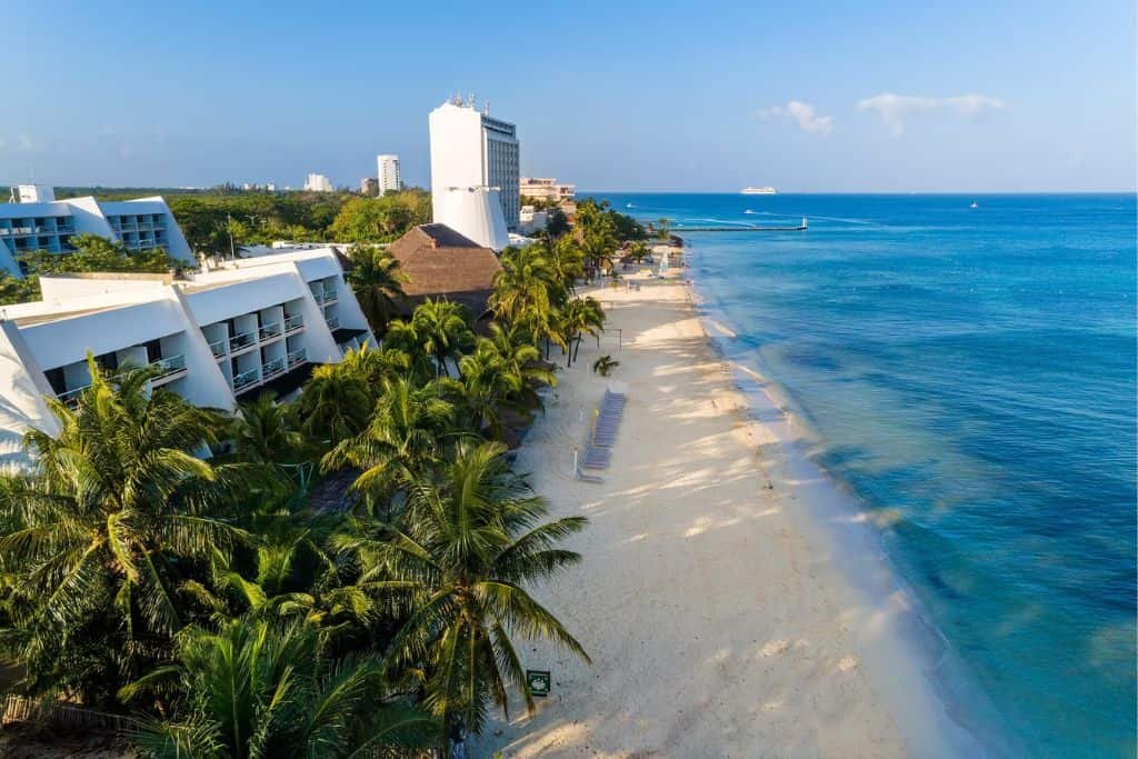 Aerial view of a white sand beach lined with palm trees and resort buildings beside clear blue ocean water. The shoreline stretches into the distance under a bright, cloudless sky.