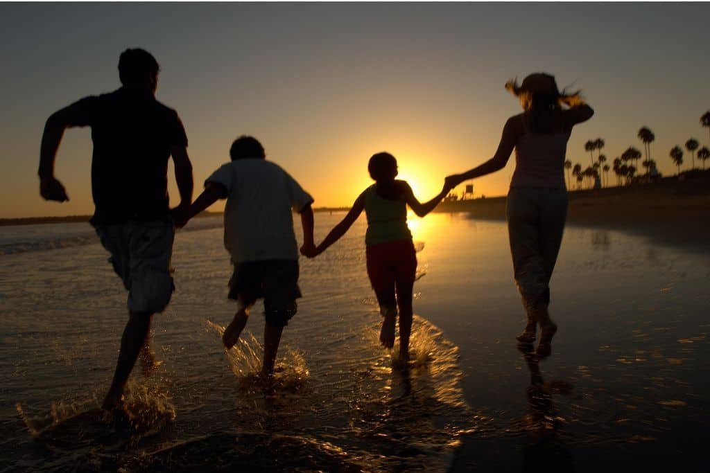 Silhouetted family of four runs hand in hand through shallow water at the beach during sunset. The golden sun glows on the horizon with palm trees faintly visible in the distance.