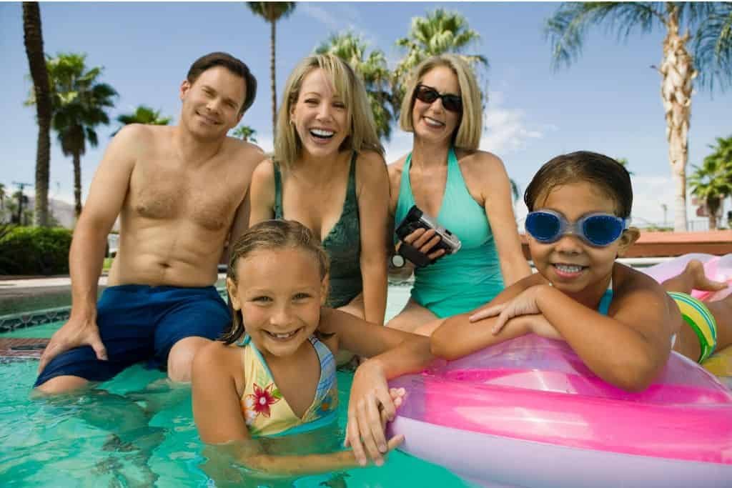 Two children play in a pool with a pink and white float while three adults smile behind them at the pool’s edge. Palm trees and clear blue sky in the background create a warm, tropical vibe.