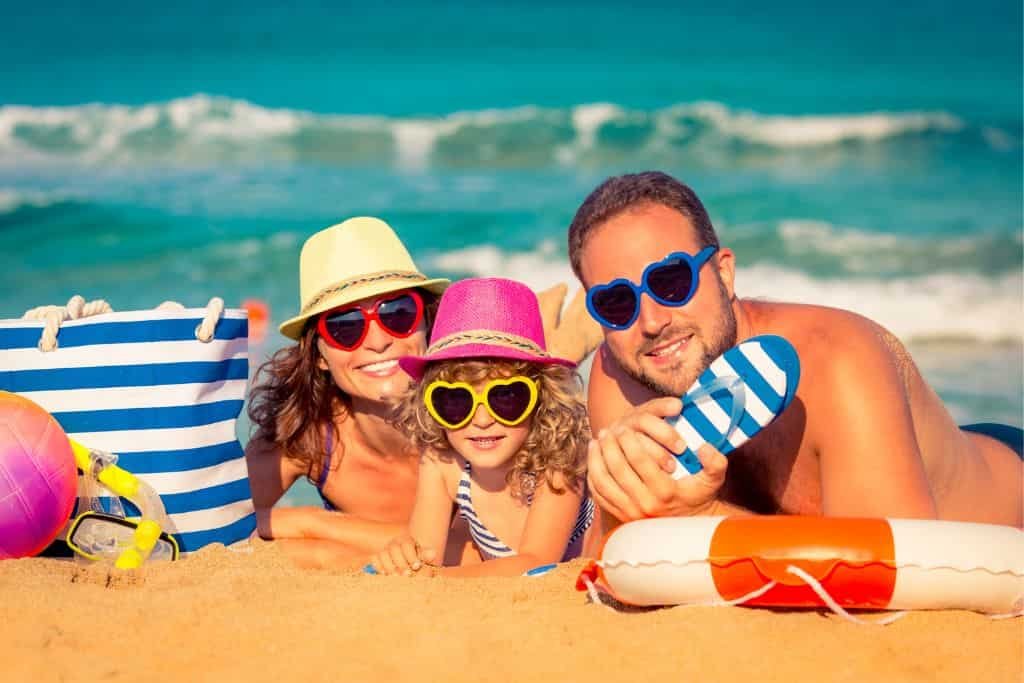 Smiling family wearing colorful hats and heart-shaped sunglasses relaxes on a sandy beach near the ocean. A striped beach bag, ball, and life preserver are placed beside them under bright sunlight.