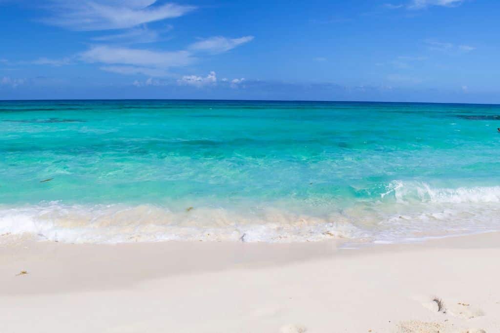 White sand beach meets clear turquoise water with small waves rolling onto the shore. The horizon blends into a bright blue sky with thin, wispy clouds.
