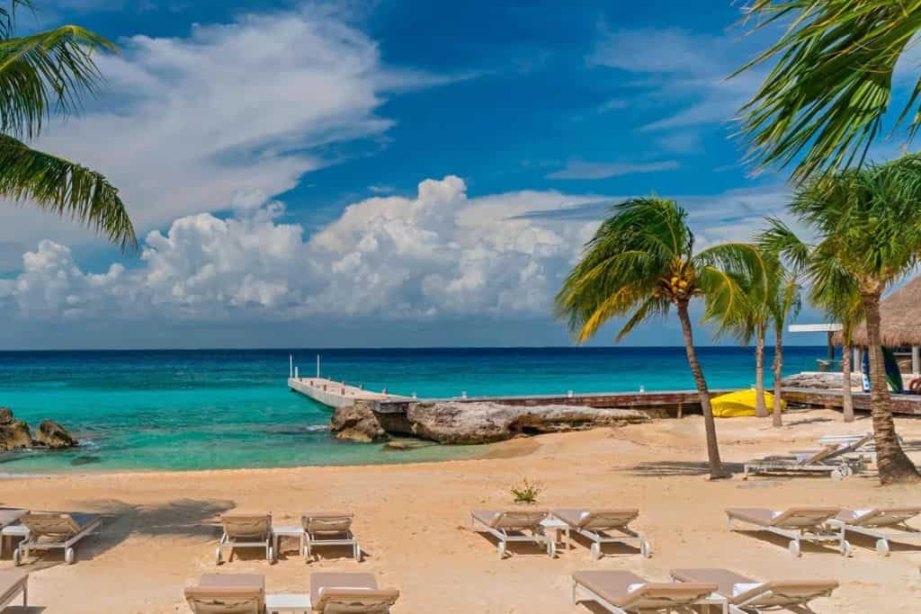 Sandy beach lined with empty lounge chairs faces turquoise ocean water and a wooden pier extending into the sea. Palm trees sway gently in the breeze under a bright blue sky with scattered white clouds.