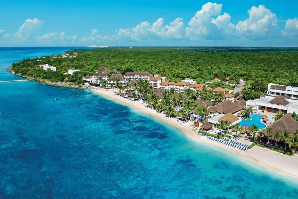 Aerial view of a tropical resort on a white sand beach with turquoise water and lush green jungle behind it. Pools, palapas, and lounge chairs line the beachfront under a bright blue sky with scattered clouds.