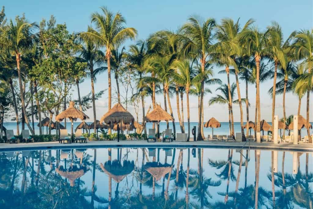 A tropical resort pool reflects tall palm trees and palapa-style umbrellas under a clear blue sky. Lounge chairs are arranged by the beach in the background, creating a relaxed vacation atmosphere.