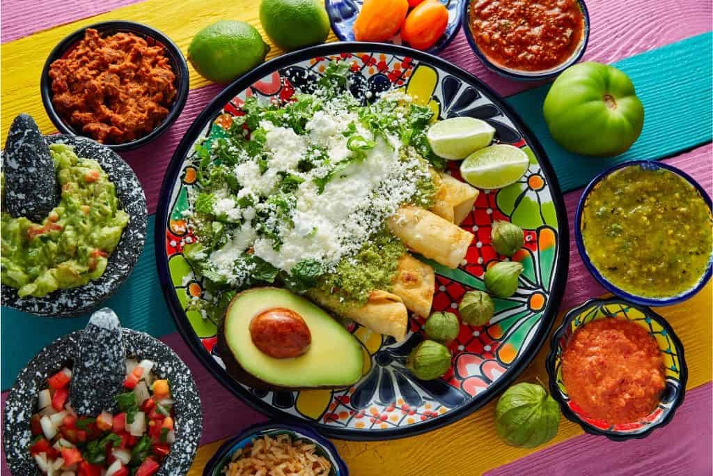 This image showcases a beautifully plated Mexican meal on a colorful, striped table. The main dish features golden, crispy tacos topped with fresh lettuce, crumbled cheese, and a drizzle of green sauce, accompanied by lime wedges and a halved avocado with a tomato in its center. Surrounding the plate are small bowls filled with guacamole, salsa, chopped vegetables, shredded meat, and a variety of sauces, creating a feast of rich textures and bold flavors.