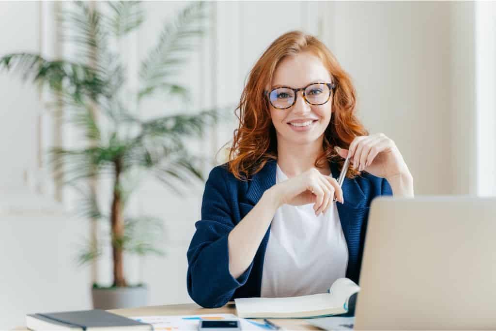 Smiling woman with red hair and glasses sits at a desk with an open laptop, notebook, and pen in hand. She’s wearing a navy blazer and white shirt, with a bright, plant-filled office space in the background.