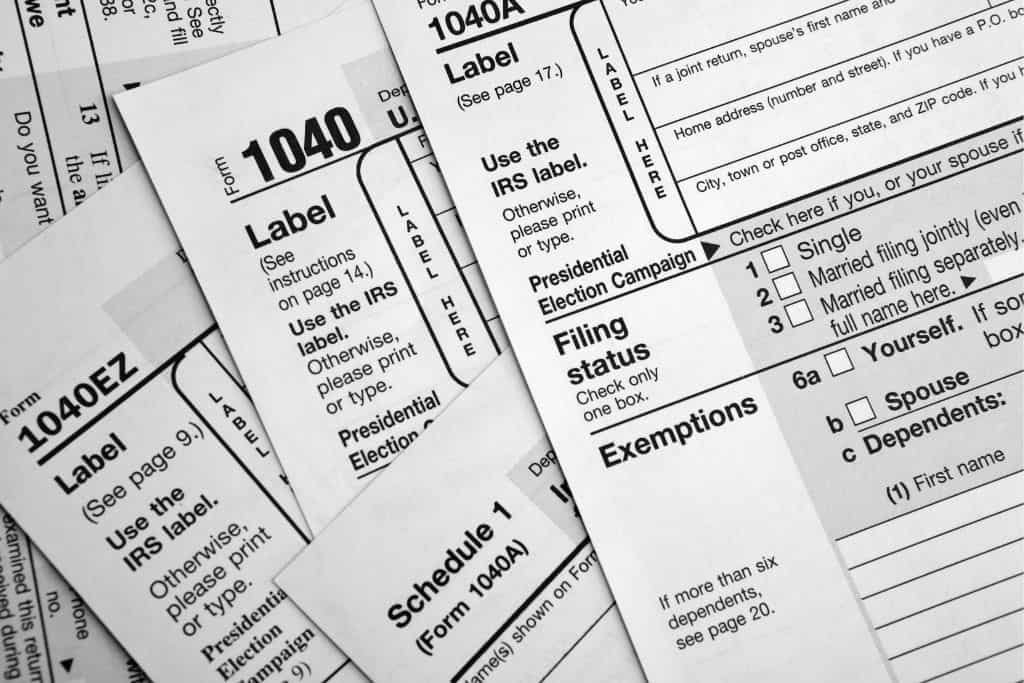 Close-up of a scattered pile of U.S. tax forms, including 1040, 1040A, 1040EZ, and Schedule 1. The black and white image emphasizes the paperwork involved in filing taxes.