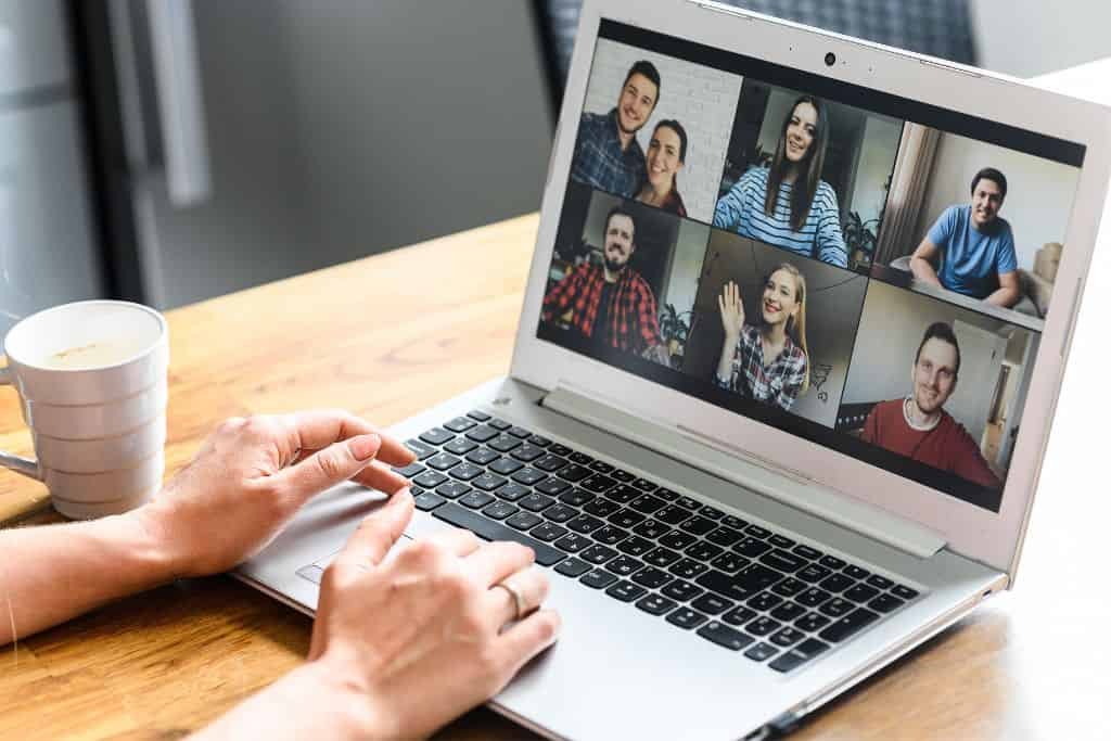 Close-up of a person typing on a laptop during a video conference with six people, each appearing in their own square on the screen. A white coffee cup sits on the wooden desk beside the laptop.