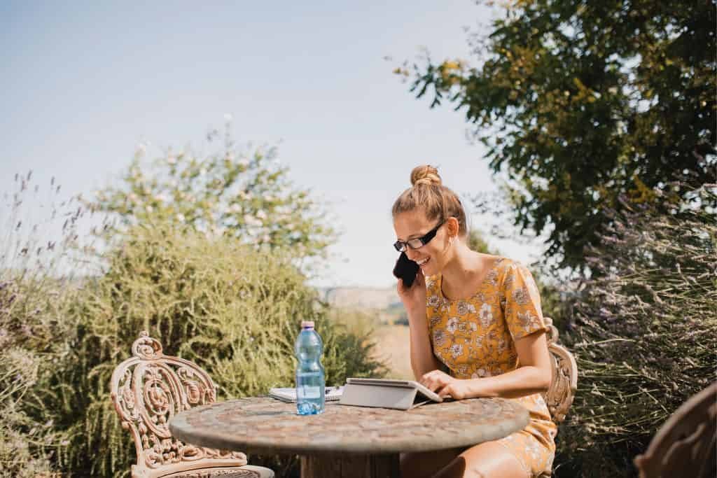 A woman in a yellow floral dress sits outdoors at a round metal table, talking on her phone while working on a tablet. She’s smiling and wearing sunglasses, with a blue water bottle on the table and lush greenery in the background.