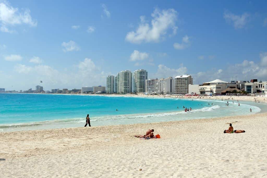 White sandy beach in Cancún with turquoise water under a blue sky, a few people lounging or walking along the shore, and high-rise hotels and resorts in the background. The scene captures a bright, sunny day with scattered clouds and calm waves.