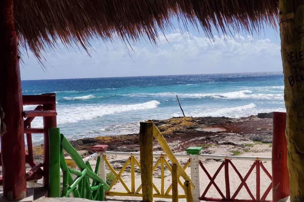 A view of the ocean from under a thatched roof, framed by colorful wooden railings painted red, yellow, and green. Waves crash gently on a rocky shoreline, and the bright blue sky meets the horizon in the distance. The scene conveys a relaxed, tropical beach vibe.