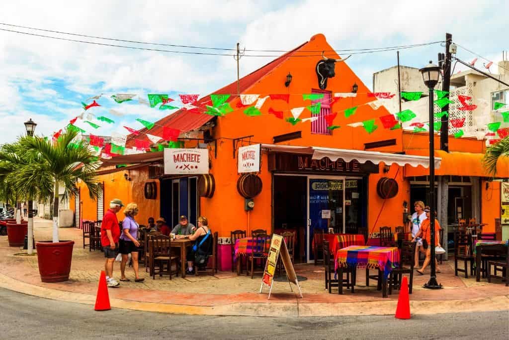 A vibrant orange Mexican restaurant with colorful papel picado flags strung overhead. People sit at outdoor tables covered in bright tablecloths, enjoying food and drinks. Palm trees and festive decor create a lively, welcoming street corner scene.