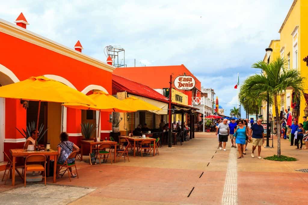 Colorful pedestrian street in Cozumel lined with vibrant buildings, outdoor restaurants with yellow umbrellas, and tourists strolling past shops and eateries under a partly cloudy sky.