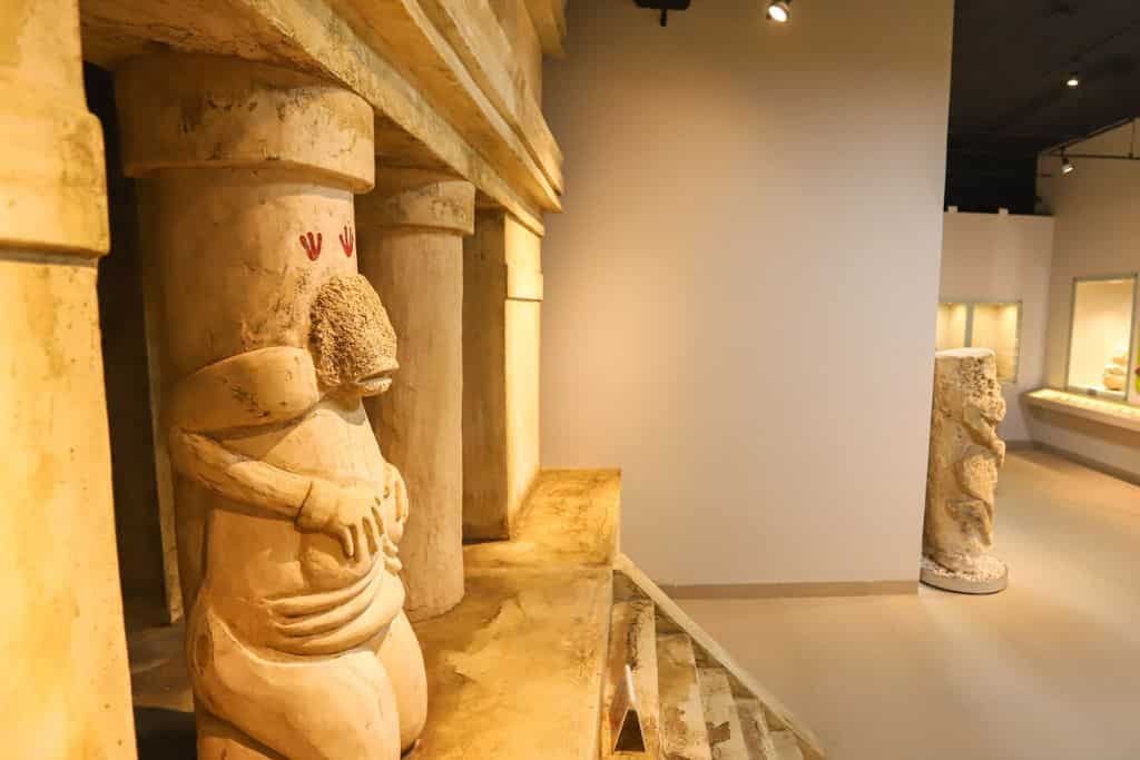 An ancient stone sculpture of a stylized human figure incorporated into a column inside a museum exhibit. The figure appears rounded with folded arms and carved details, standing beneath a decorative lintel. Soft lighting highlights the warm tones of the stone and the architectural details on display.