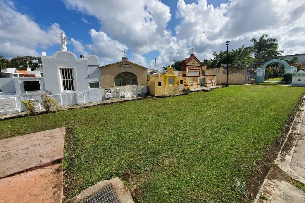 A cemetery with brightly colored mausoleums in white, yellow, and peach tones lining a neatly kept grassy path. Each tomb has unique architectural details, some with crosses and statues on top. The sky above is blue with large white clouds, creating a peaceful setting.