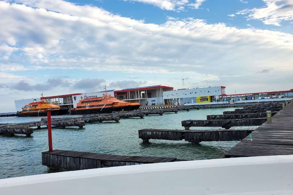 A waterfront with empty wooden docks leading out into turquoise water. In the background, bright orange ferries are moored near a terminal building with a red roof. The sky is partly cloudy, creating a peaceful coastal harbor scene.