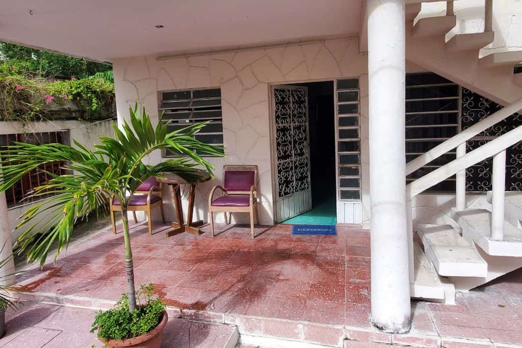 A small outdoor patio area with two purple chairs and a round table placed near a doorway. A potted palm tree adds greenery beside the tiled red floor, and a white staircase curves upward to a second level. The open door reveals a glimpse of the interior hallway.