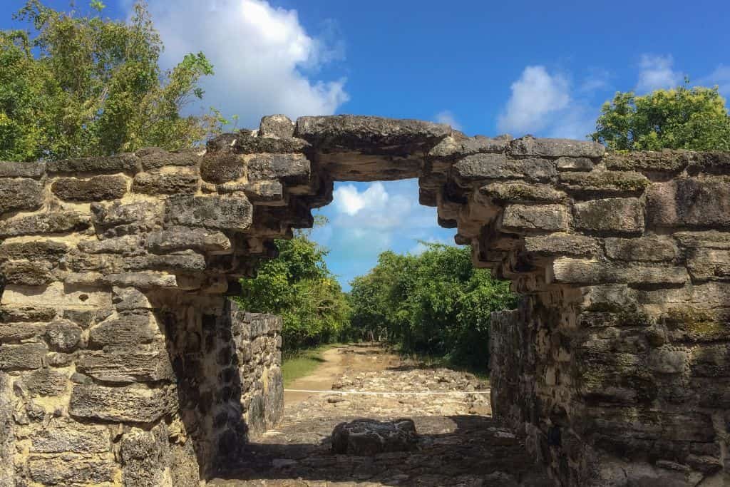 A stone archway from ancient ruins, surrounded by lush green vegetation under a bright blue sky. The weathered stones form a pathway leading deeper into the site, suggesting historical significance and architectural remnants of a past civilization. The open arch frames the natural scenery beyond.