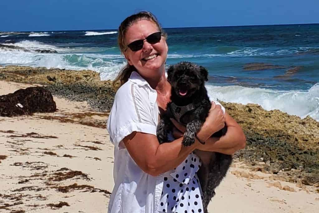 A smiling woman wearing sunglasses holding a small black dog on a sandy beach. Waves crash onto the rocky shoreline in the background, and the bright blue sky enhances the tropical setting. Both the woman and the dog appear relaxed and happy by the ocean.