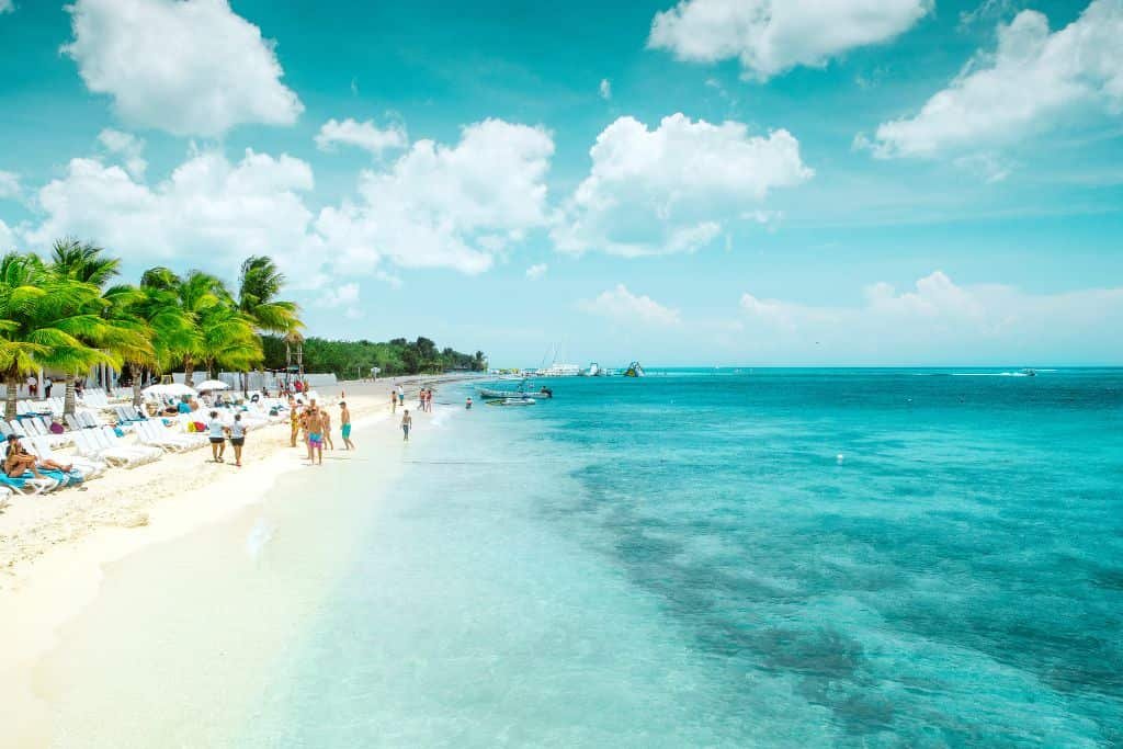 Idyllic tropical beach with white sand, turquoise waters, palm trees, and vacationers lounging or strolling along the shoreline under a bright blue sky with fluffy clouds.