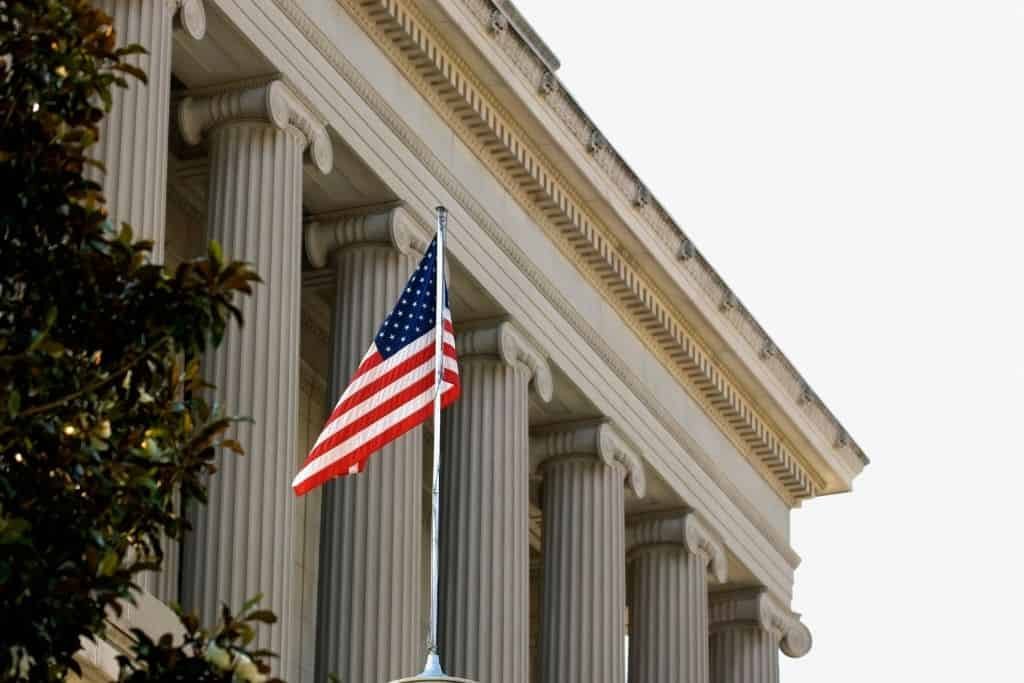 American flag flying in front of a classical U.S. government building with tall stone columns and ornate architectural details, partially framed by leafy tree branches.