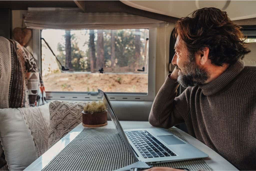 A man sitting inside a cozy camper van, working on a laptop while gazing out the window at a peaceful forest scene. He is dressed warmly in a sweater, and the interior is decorated with soft cushions and natural tones, creating a comfortable and serene atmosphere that blends work with outdoor adventure.