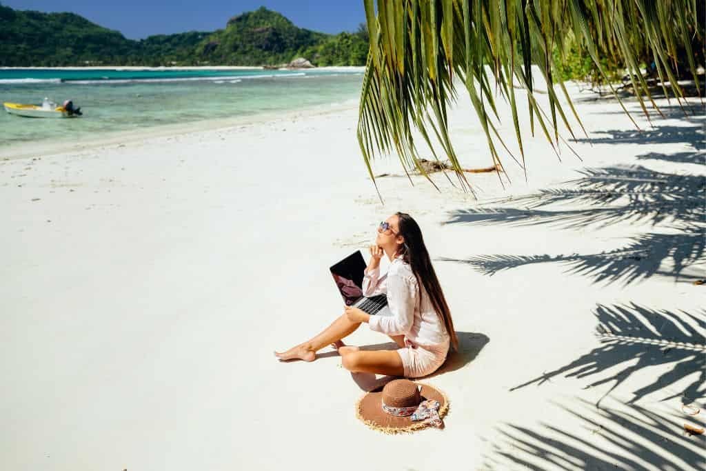 A woman sitting on a sandy beach, holding a laptop while gazing toward the ocean. She sits next to a straw hat under the shade of palm trees, with a bright and tranquil tropical backdrop of clear blue water and distant green hills. The scene captures a serene moment of remote work by the sea.