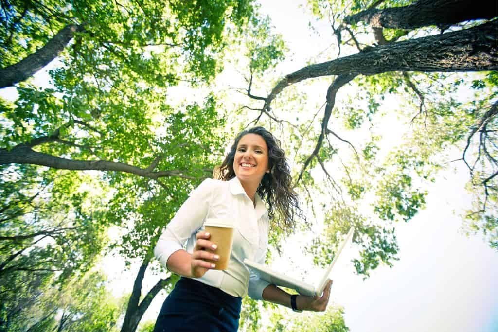 A woman standing outdoors under tall, leafy trees. She is holding a laptop in one hand and a takeaway coffee cup in the other, smiling as she looks off into the distance.