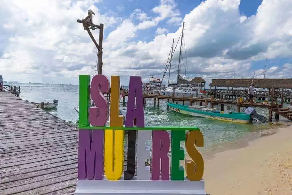 A colorful "Isla Mujeres" sign in bright green, purple, yellow, and pink letters on a sandy beach. In the background, there are wooden docks with boats and yachts anchored in calm turquoise water, set under a partly cloudy sky. The scene captures a lively marina atmosphere on Isla Mujeres, Mexico.