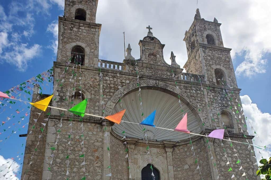 A stone church in Merida with twin bell towers rises against a partly cloudy sky, decorated with colorful triangular pennant flags. The building’s arched entrance and cross detail highlight its historic architecture.
