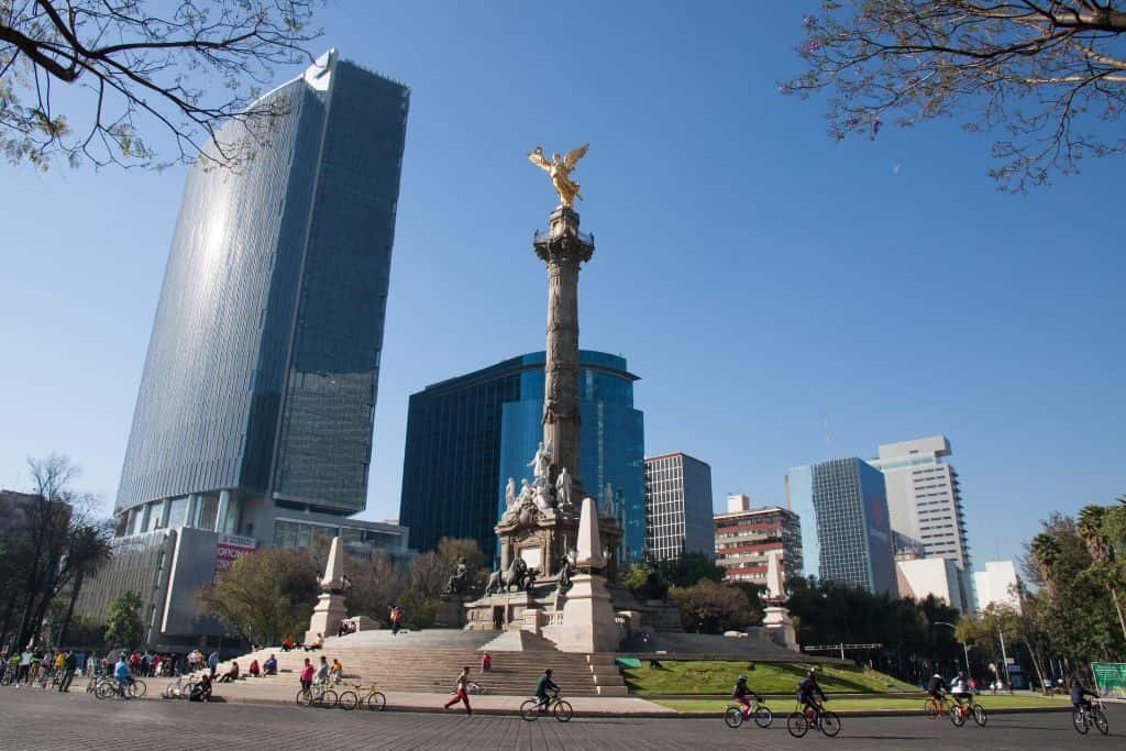 A tall monument topped with a golden angel stands in a busy city roundabout surrounded by modern skyscrapers. People walk and cycle around the landmark under a clear blue sky.