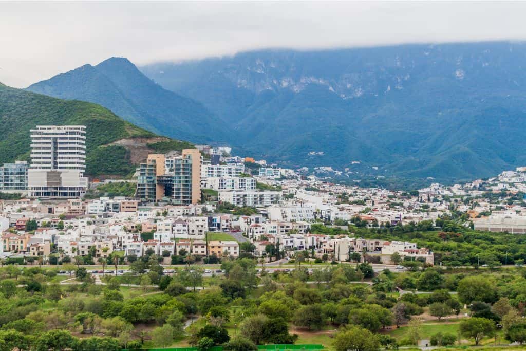 A modern city with white buildings and tall towers is set against lush green mountains. Low clouds hover over the peaks, and dense vegetation surrounds the urban area.
