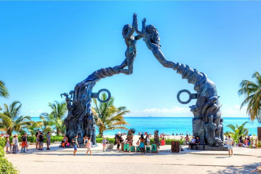 A large beachfront sculpture of two figures reaching toward each other forms an archway with ocean views behind it. Palm trees and people walking along the promenade add to the lively tropical scene.
