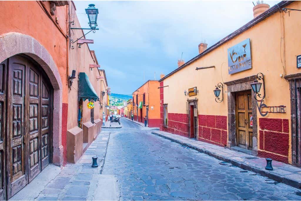 A narrow cobblestone street is lined with colorful colonial-style buildings in shades of orange and red. Wooden doors, lanterns, and wrought-iron accents add charm to the quiet historic scene.