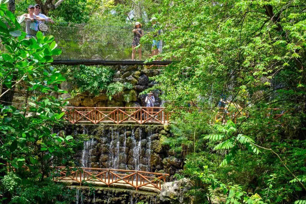 A lush, green aviary at Xcaret, featuring cascading waterfalls flowing over rocky terraces with wooden bridges and walkways. Visitors stand on an elevated glass bridge, observing the vibrant tropical environment filled with dense foliage. Sunlight filters through the trees, creating a serene and immersive jungle-like atmosphere.