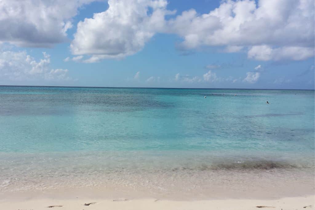 A tranquil Caribbean beach with soft white sand leading to crystal-clear turquoise waters. Gentle waves lap at the shore under a partly cloudy sky, creating a peaceful and inviting tropical scene. The calm ocean stretches to the horizon, evoking a sense of relaxation and escape.