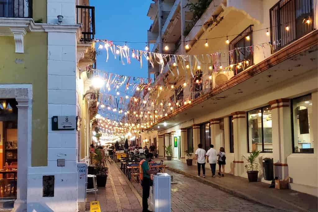 This image captures a charming evening scene on a narrow cobblestone street lined with colonial-style buildings. String lights and festive banners hang overhead, casting a warm glow as people stroll and enjoy the ambiance.